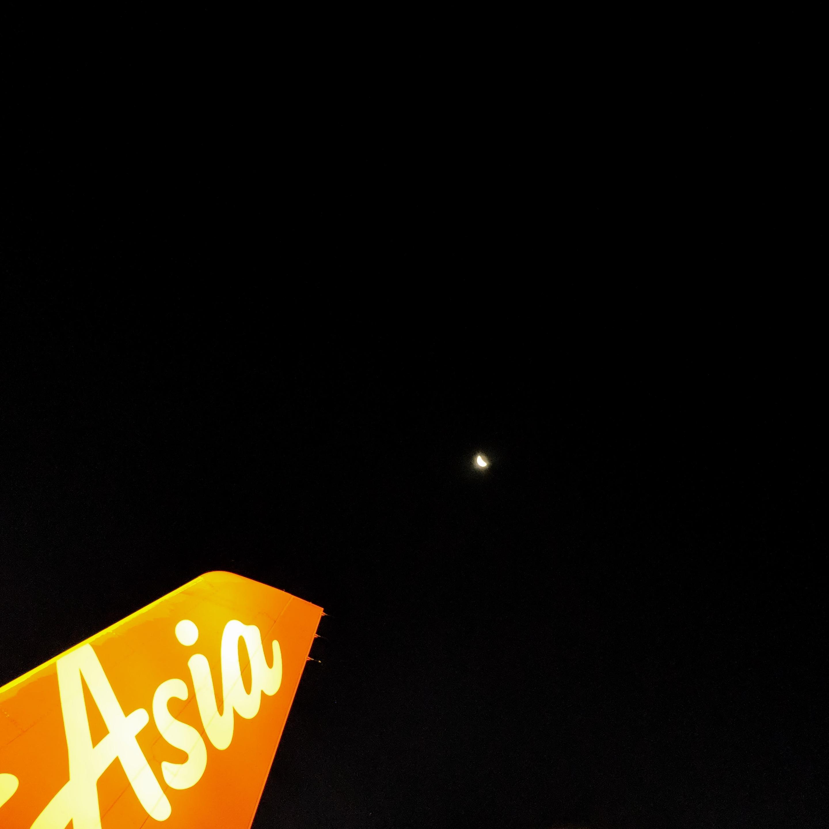 Photo of an airplane's tail wing with "Asia" on it pointing to the moon in the night sky.